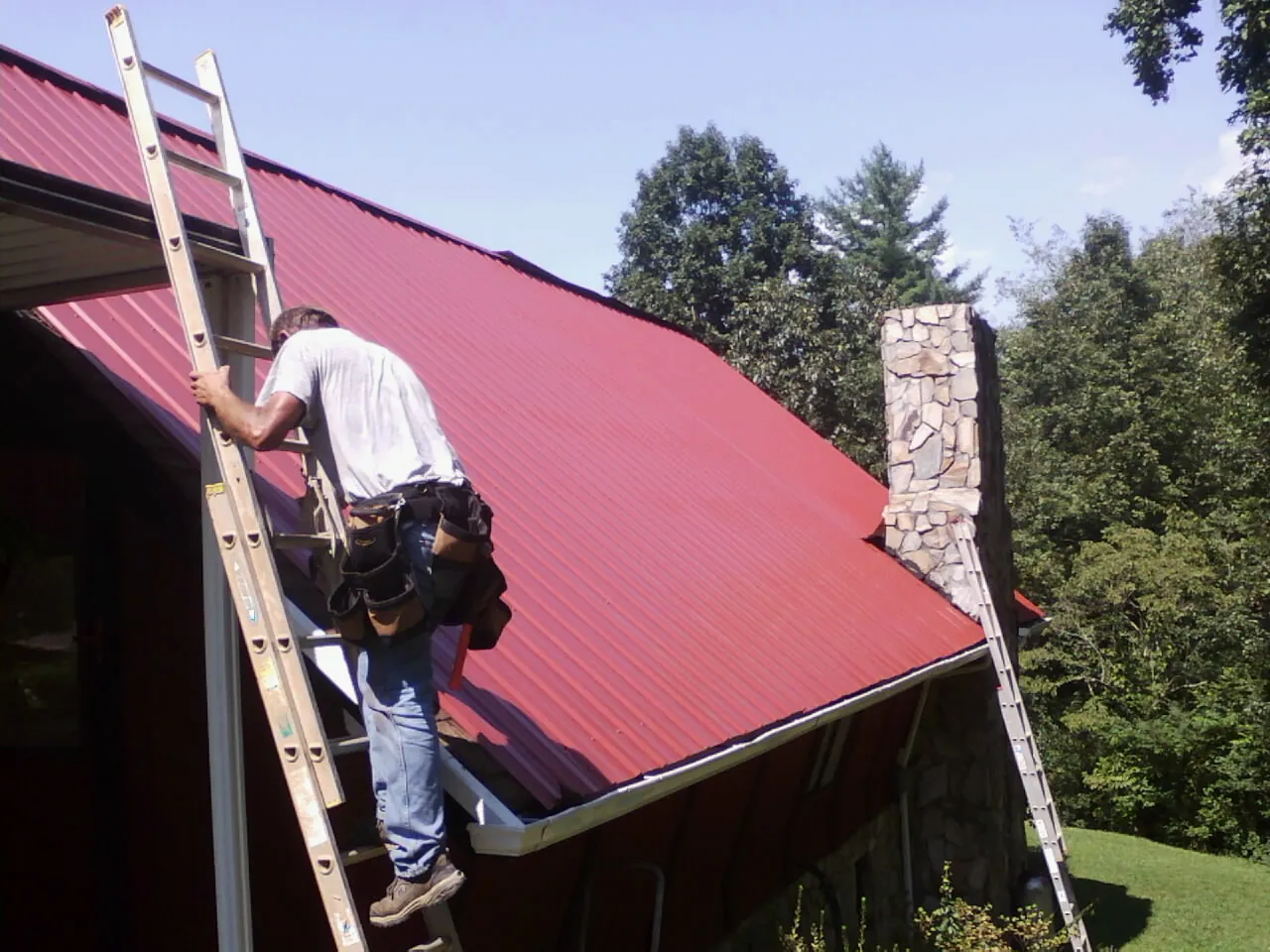 Roofer on a ladder beside a red metal roof and stone chimney — Soffit & Fascia Repair by Roofing Brothers in Reading